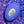 A blue oval personal massager from We-Vibe with a silver metallic center plate, covered in water droplets, set against a background of purple and blue rippling water.
