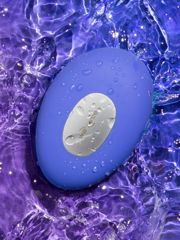 A blue oval personal massager from We-Vibe with a silver metallic center plate, covered in water droplets, set against a background of purple and blue rippling water.