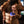 A shirtless man holding a blue circular personal massager against his lower torso, with water droplets spraying in the air and a dark, moody background.
