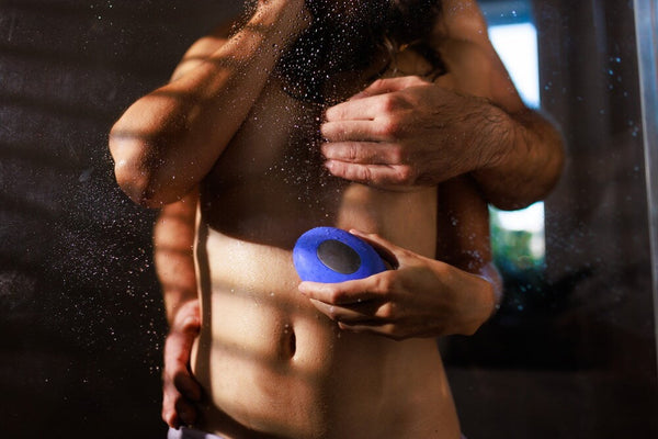 A shirtless man holding a blue circular personal massager against his lower torso, with water droplets spraying in the air and a dark, moody background.