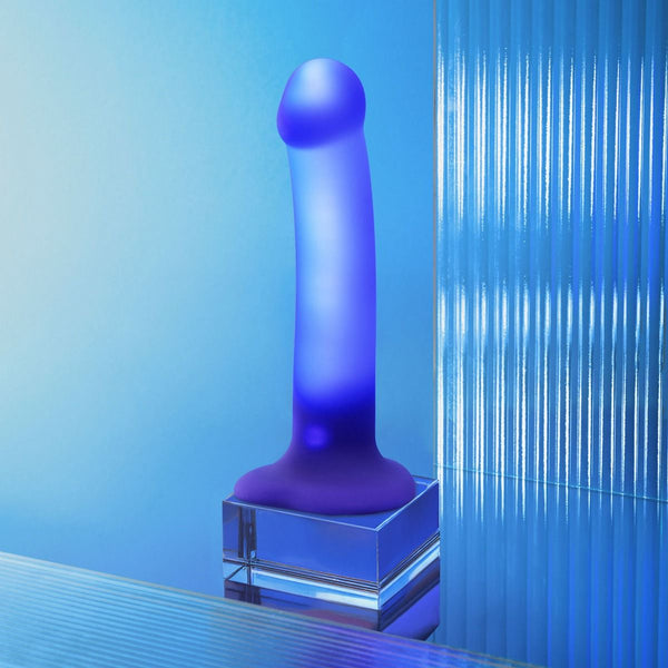 Blue dildo from Strap-on-Me with internal LED lights on a reflective surface with a gradient blue background
