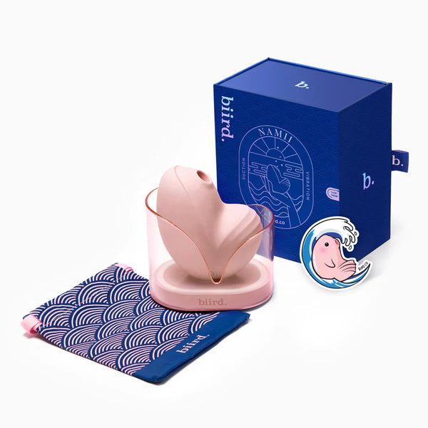 Biird Namii Rechargeable Dual "Sucker" & Vibrator in cream. Shown in its cream hued holder alongside its navy blue packaging and other Biird paraphernalia..