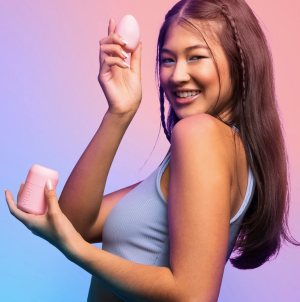 Smiling female model in lilac top holding up the VUSH Plump Rechargeable Palm Vibrator in light pink alongside its cap and case.