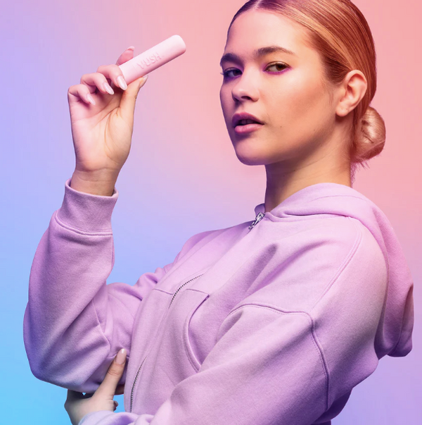 Female model wearing a lilac top holding up a VUSH Gloss Rechargeable Bullet Vibrator in light pink with its cap on.