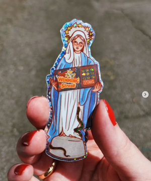 Hand with red nail polish holding a sticker of 'Our Lady of Eternal Fortune', which is an angel in Blue and White holding a winning lottery ticket.
