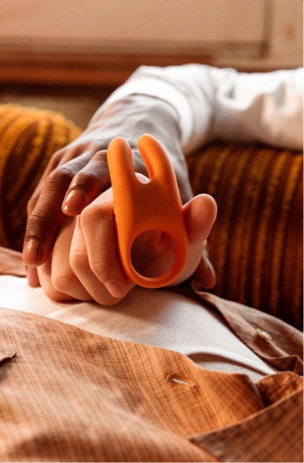 Two people holding hands outstretched on a couch with one of them holding the Vush Orb cock ring in Orange.