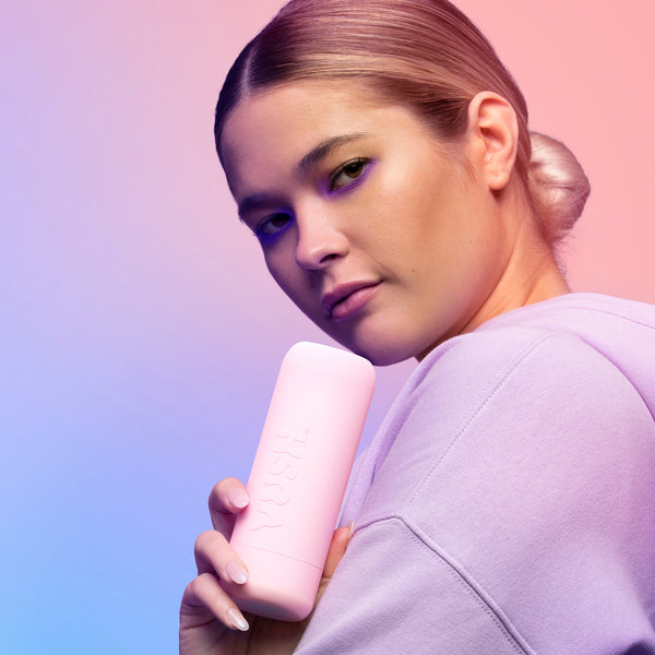 Woman wearing a purple top holding up the Vush Glow Rechargeable Mini Rabbit Vibrator in pink, in its case.