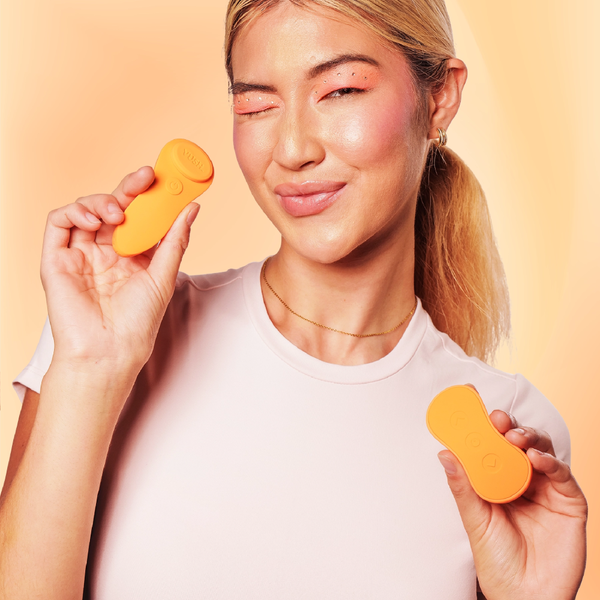 Winking woman in a white t-shirt holding up the Vush Luna Remote Controlled Wearable Vibrator in orange, with its remote control in the other hand.