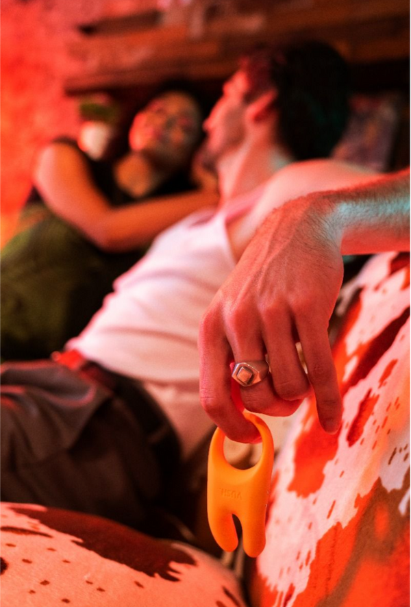 Artsy picture of a couple sat on the couch with one dangling the Vush Orb cock ring in Orange from their hand.