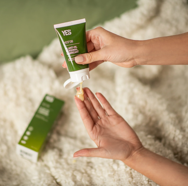 A woman's hands squeezing a small bottle of Yes OB plant oil based personal lubricant onto the pads of her fingers. Background of a white fluffy blanket on a green surrounding.