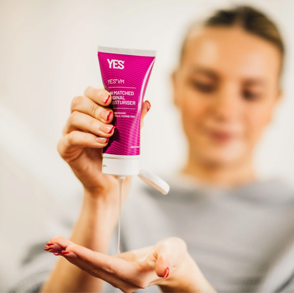 A slightly blurred menopausal woman squeezes a purple bottle of Yes VM vaginal moisturizer into her hand.