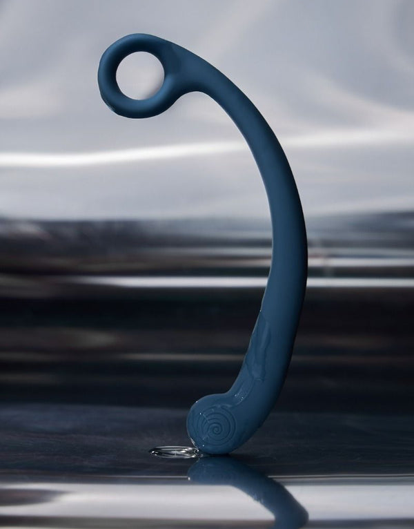 The Svibe Axi vibrator stands on one end dripping with lubricant in front of a blurry background of a lake. 