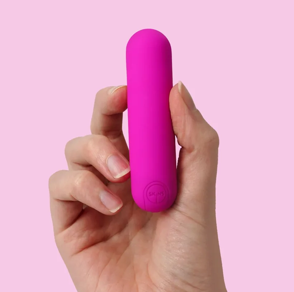 Hand holding up a pink Skins Super Excite Rechargeable Micro Vibrator Shown on a light pink background.