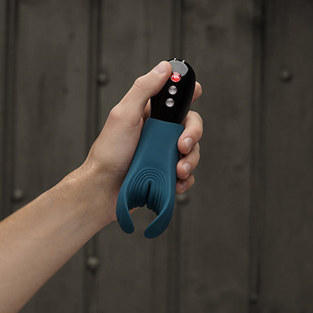 Mood photo of a hand holding a turquoise Fun Factory Manta blow job vibrator for couples. Toy has a black handle, and background is of a fence - Sex Siopa, Ireland's favourite sex toy shop