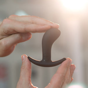 Hands holding the Fun Factory Bootie silicone butt plug sex toy in black. Sunlit hewn background.